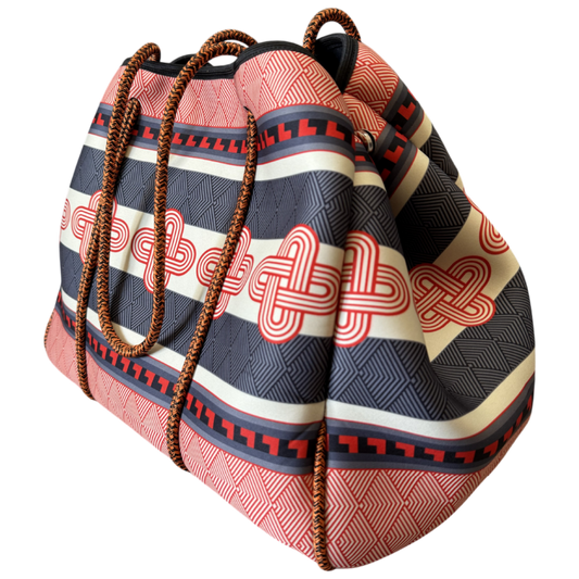 A patterned bucket-style tote bag with woven brown rope handles, featuring horizontal stripes in red, black, and cream with red geometric and knot-like motifs, shown on a plain white background.