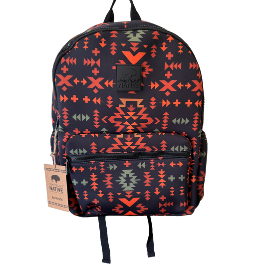 A backpack with a dark navy blue base featuring a vibrant geometric Southwestern pattern in orange, red, and sage green. It has a front zippered pocket, black top handles, and a hanging paper tag on the left side.