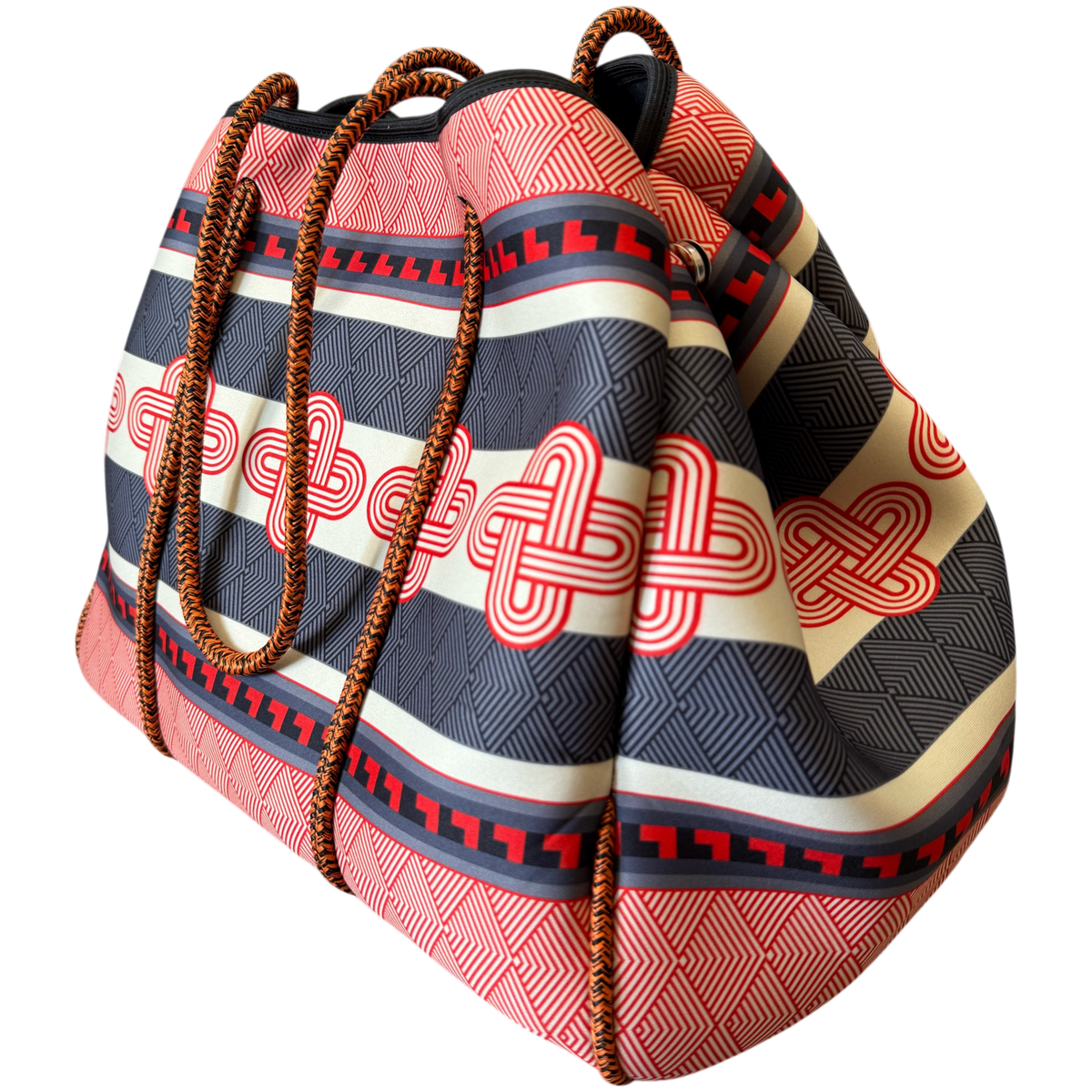 A large drawstring tote bag with a bold geometric pattern in coral pink, navy blue, cream, and red, featuring brown braided rope handles, shown on a plain white background.