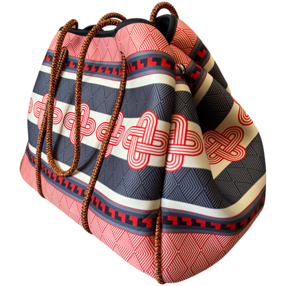 A large drawstring tote bag with a bold geometric pattern in coral pink, navy blue, cream, and red, featuring brown braided rope handles, shown on a plain white background.