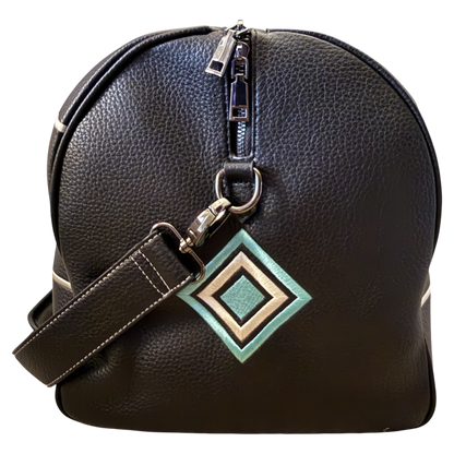A black pebbled leather duffel bag with a silver zipper, a matching shoulder strap, and a geometric diamond-shaped metal charm in green, beige, and white, shown against a plain white background.