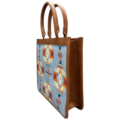 A structured tote bag with brown faux-leather handles and side panels, featuring a light blue front panel decorated with a Southwestern geometric pattern in orange, red, yellow, and dark blue.
