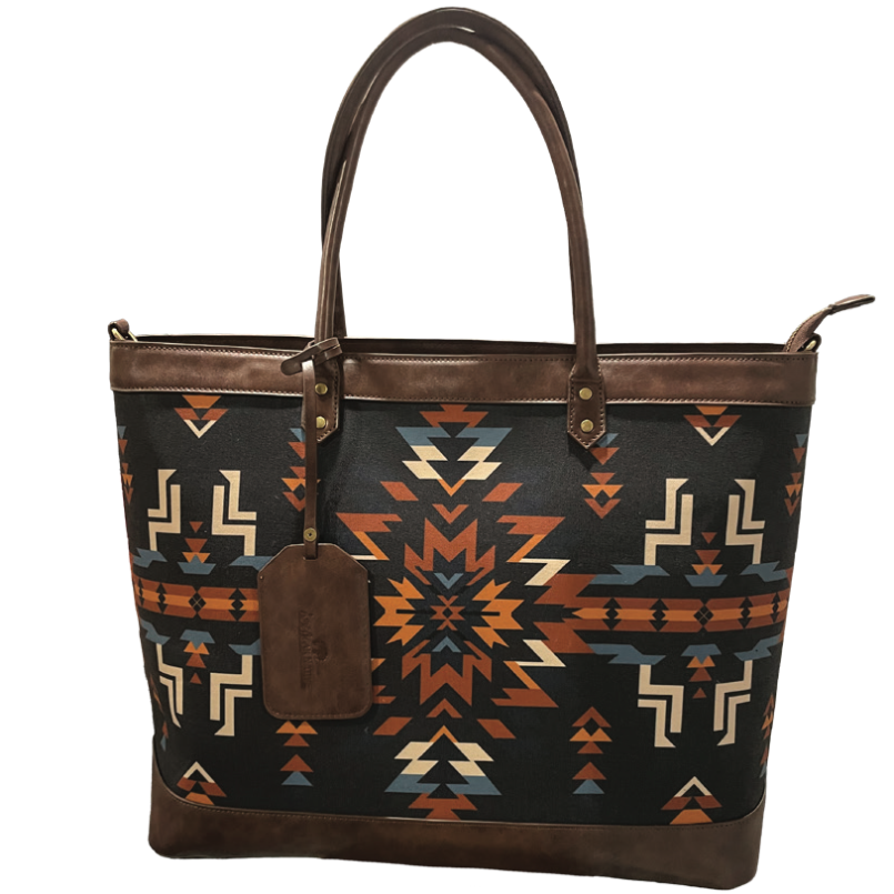 A black canvas tote bag with a brown leather trim and handles, featuring a Southwestern geometric pattern in orange, beige, and blue. A brown leather hangtag is attached to the front.