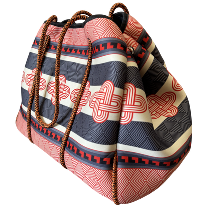 A patterned bucket-style tote bag with woven brown rope handles, featuring horizontal stripes in red, black, and cream with red geometric and knot-like motifs, shown on a plain white background.