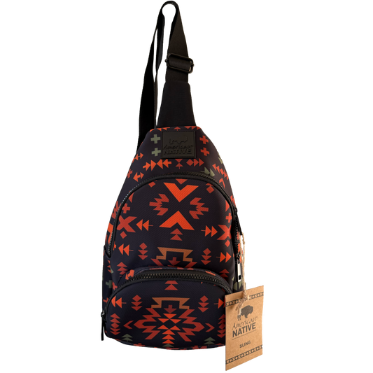 A small sling backpack with a black base and a Southwestern geometric pattern in orange, red, and brown. It features a black adjustable shoulder strap, a front zippered pocket, and a hanging paper tag that reads 'American Native'.