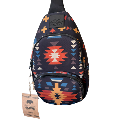 A sling backpack with a Southwestern geometric pattern in navy, red, orange, yellow, and white, featuring a black brand patch and a hanging paper tag.