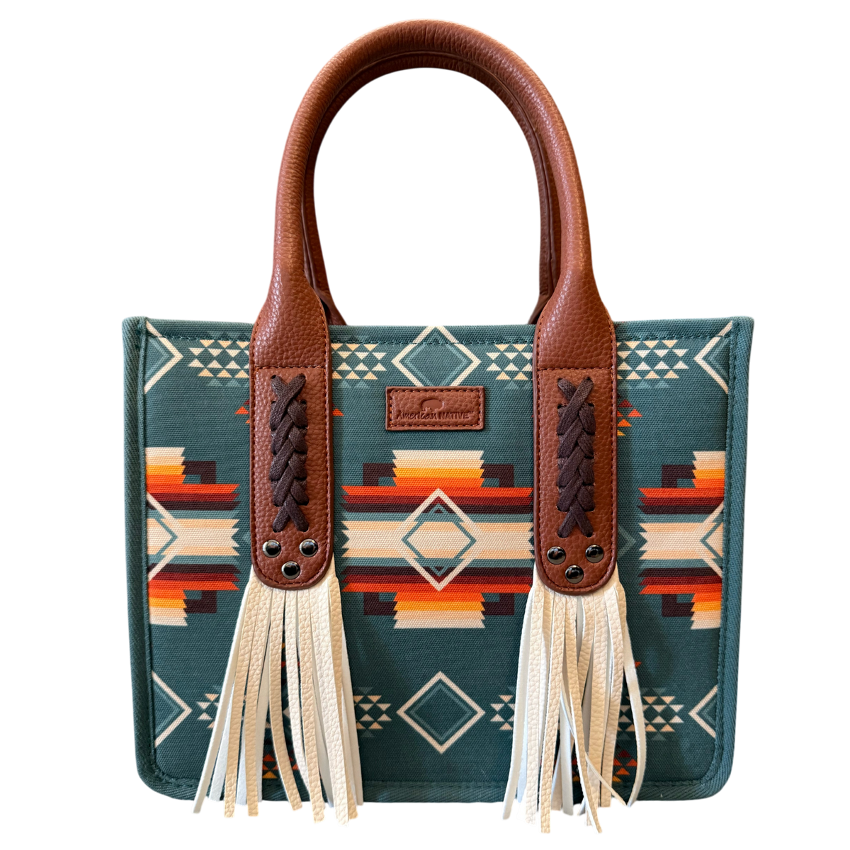 A structured tote bag with a Southwestern geometric pattern in teal, brown, orange, and cream, featuring brown faux-leather handles and long cream tassels.