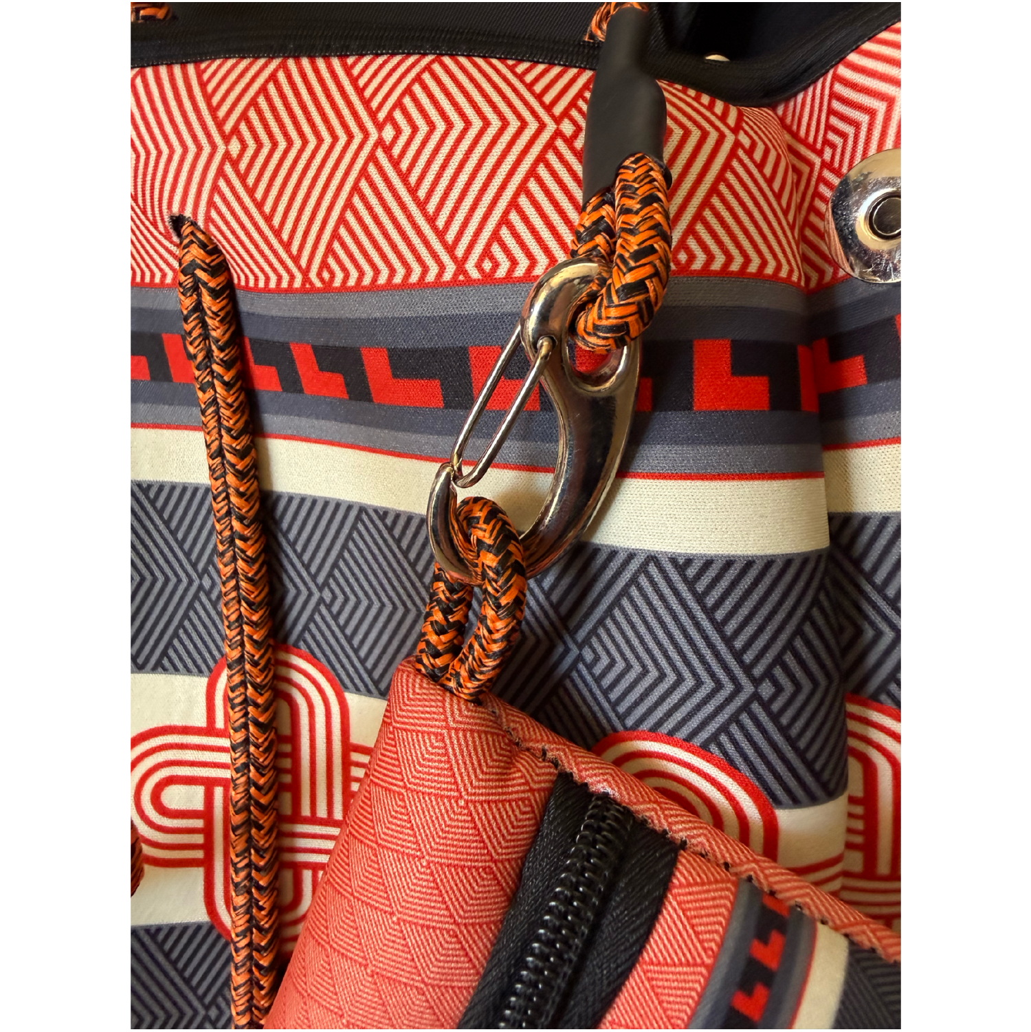 Close-up detail of a patterned backpack showing orange and black braided webbing straps, a silver metal carabiner, and a geometric print in red, black, gray, and cream.