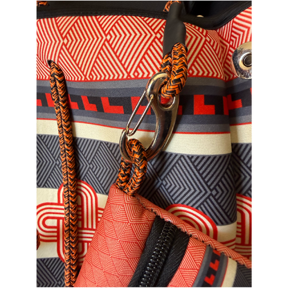 Close-up detail of a patterned backpack showing orange and black braided webbing straps, a silver metal carabiner, and a geometric print in red, black, gray, and cream.